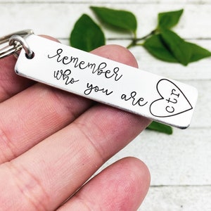 Puede incluir: Un llavero rectangular plateado con las palabras "remember who you are" en escritura cursiva. Un corazón con las letras "ctr" en su interior está a la derecha. El llavero está sostenido en una mano.