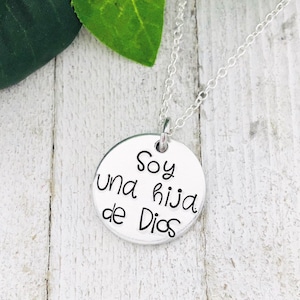 Könnte beinhalten: Eine silberfarbene Halskette mit Anhänger und einem runden Charm. Auf dem Charm steht der Schriftzug "Soy una hija de Dios" in schwarzer Schrift. Die Kette ist ebenfalls silberfarben. Der Hintergrund ist weißes Holz.