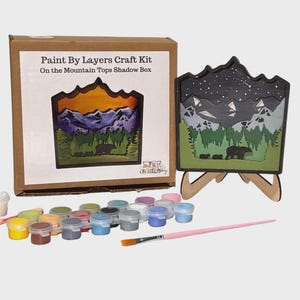 May include: A Paint By Layers Craft Kit with a shadow box featuring a mountain scene with bears. The kit includes a box, paint pots, a paintbrush, and a wooden easel. The box reads "Paint By Layers Craft Kit On the Mountain Tops Shadow Box."