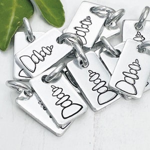 May include: A collection of rectangular silver-toned metal charms, each featuring a jump ring at the top. The surface of every charm is engraved with a black line drawing of a stack of rounded stones, resembling a cairn or zen stone tower.