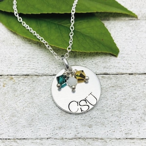 May include: A silver pendant necklace with a chain. The pendant is a round, brushed metal disc with the letters "CSU" engraved on it. Three colorful crystals are attached to the pendant. The necklace is displayed on a white wooden surface.