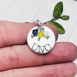 May include: A silver pendant necklace with the letters "UNC" stamped on a round disc. The pendant features three colorful crystal charms in blue, yellow, and white. The necklace is held in a hand.