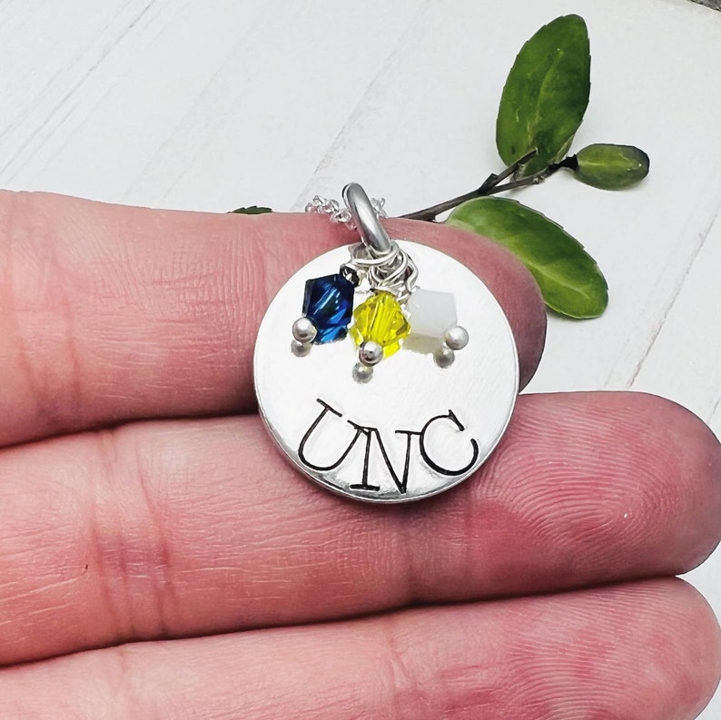 May include: A silver pendant necklace with the letters "UNC" stamped on a round disc. The pendant features three colorful crystal charms in blue, yellow, and white. The necklace is held in a hand.