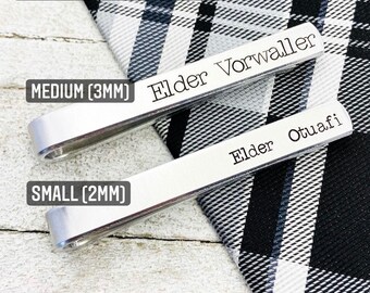 May include: Two silver tie clips with different font sizes. The larger tie clip reads "Elder Vorwaller" in a 3mm font. The smaller tie clip reads "Elder Otuafi" in a 2mm font. The tie clips are on a black and white checked fabric.