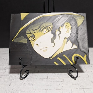 May include: A black and yellow illustration of a character with a hat, displayed on a black easel. The artwork features a detailed face with large eyes and a focused expression. The color palette is primarily black and yellow.