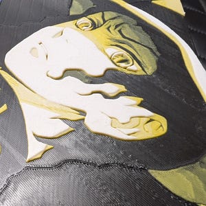 May include: A 3D-printed artwork depicting a stylized face with white and yellow details on a black background. The piece has a textured surface, resembling a relief sculpture.
