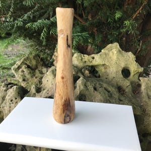 May include: A handcrafted wooden vase with a natural, light brown finish. The vase has a slender, elongated shape with a slightly flared base and a smooth, polished surface. The vase is displayed on a white surface against a backdrop of greenery and rocks.