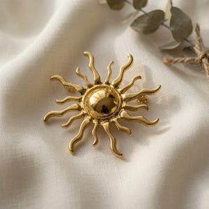 May include: A gold-toned metal brooch shaped like a sun with a smooth, rounded center and multiple radiating, wavy rays extending outward. The piece features a polished, reflective surface that catches the light, creating a bright, metallic appearance.