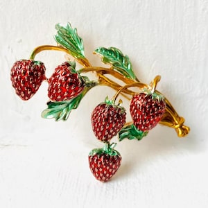 May include: A gold-tone metal brooch featuring four red enamelled strawberries with textured surfaces. The strawberries are attached to gold stems with green enamelled leaves, creating a cluster design that mimics a small branch of fruit.