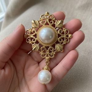 May include: A gold-tone brooch featuring an intricate, openwork filigree frame in a square shape with rounded corners. A large, round, white faux pearl is set in the center, and a smaller, round, white faux pearl hangs from the bottom of the frame.