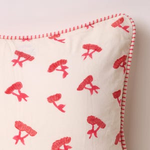 May include: Cream-colored square pillow with a repeating pattern of red floral embroidery. The pillow has a red and white striped border, adding a decorative touch. The floral design gives the pillow a fresh, spring-like feel.