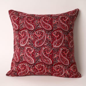 Puede incluir: Un cojín cuadrado decorativo con un estampado de cachemira rojo. El cojín presenta un diseño repetido de grandes formas de cachemira estilizadas en tonos de rojo, blanco y negro. El cojín es probablemente de tela y está diseñado para la decoración del hogar.