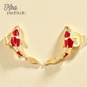 May include: Gold-coloured koi fish earrings with red enamel accents and small clear stones. The earrings are shaped like a koi fish. The Kira JewelStudio logo is in the upper left corner.