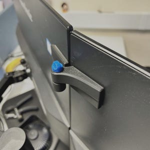 Puede incluir: Un soporte de alineación de monitores de plástico negro texturizado con una perilla azul brillante en la parte superior. El dispositivo está sujeto al lateral de dos monitores de computadora negros adyacentes para mantenerlos alineados.