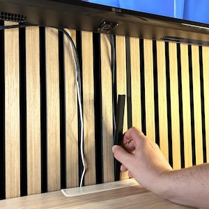May include: A wooden slat wall panel with black vertical gaps. A hand is holding a black cable management strip. White and black cables are visible. The panel is above a light-colored wooden shelf.