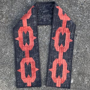 Pode incluir: Um cachecol acolchoado com um design geométrico. O cachecol apresenta um fundo preto com um padrão repetitivo de formas vermelhas. O cachecol é feito de tecido e tem uma forma retangular. O cachecol é provavelmente feito à mão e adequado para o clima frio.