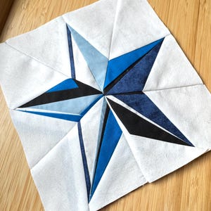 May include: A square, white fabric block with a blue star design. The star features shades of blue and black, with a geometric, angular pattern. The block is likely part of a larger quilt or textile project.