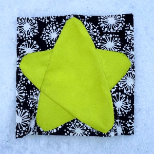 Puede incluir: Una aplicación de estrella verde lima está centrada en un cuadrado de tela estampada en blanco y negro. La tela tiene un patrón repetitivo de explosiones de estrellas blancas sobre un fondo negro. La estrella está hecha de un material suave y texturizado.