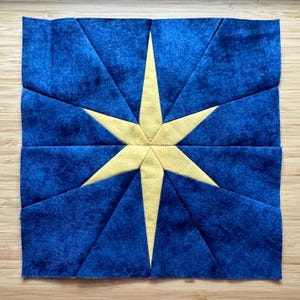 Puede incluir: Un trozo de tela cuadrado con un diseño de estrella amarilla sobre un fondo azul. La estrella está centrada, con puntas que se extienden hasta los bordes. La tela tiene una apariencia texturizada, lo que sugiere una construcción acolchada o de patchwork.