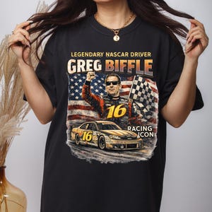 May include: Black t-shirt with a graphic of NASCAR driver Greg Biffle. The text "LEGENDARY NASCAR DRIVER GREG BIFFLE" is above an image of the driver and his race car. The American flag and a checkered flag are also in the graphic.