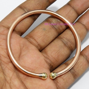 May include: A rose gold-colored bangle bracelet with gold-colored ball accents at the ends. The bracelet is circular and appears to be made of metal. The ends of the bracelet have a curved design.