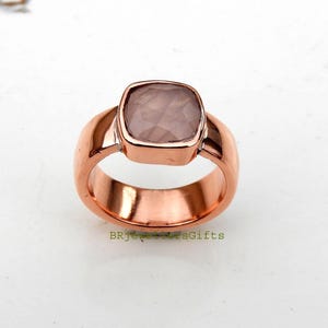 Könnte beinhalten: Ein kupferfarbener Ring mit einem quadratischen, facettierten, blassrosa Edelstein in einer glatten Metallschiene. Der Text BRJewellersGifts ist in kleinen grünen Buchstaben am unteren Rand des Rings mit polierter, reflektierender Oberfläche gedruckt.