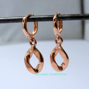 Könnte beinhalten: Ein Paar roségoldene Creolen mit einem verdrehten Tropfendesign. Die Ohrringe sind an kleinen Reifen aufgehängt und haben eine polierte Oberfläche. Die Ohrringe werden vor einem hellen Hintergrund präsentiert.