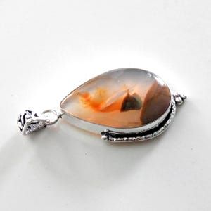 May include: A teardrop-shaped pendant with a silver setting. The pendant features a polished stone with orange, brown, and gray hues. The silver setting has decorative details and a loop for a chain.
