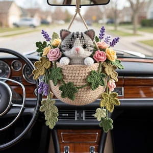Op de afbeelding: Een gehaakte kat in een geweven mand, versierd met kleurrijke bloemen en bladeren, hangt aan de achteruitkijkspiegel van een auto. De kat heeft grijs-witte strepen, met roze accenten. De mand is beige en de bloemen zijn roze, geel en paars.