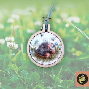 May include: A small circular wooden embroidery hoop containing a cross-stitch design of a dark gray mole with a pink nose emerging from brown earth surrounded by green grass, blue flowers, and two small bees, suspended by a light blue string.