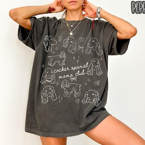 May include: Dark grey t-shirt featuring white line drawings of Cocker Spaniels and the text "cocker spaniel mama club". The shirt has a relaxed fit and is made of a soft material. The shirt is a comfortable and stylish choice for dog lovers.