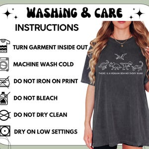 May include: A charcoal gray t-shirt featuring a white line-art graphic of four dinosaurs and the text "THERE IS A REASON BEHIND EVERY ROAR." The image includes a washing and care instruction chart with icons for turning inside out, cold wash, no bleach, no dry clean, low heat dry, and no ironing on the print.