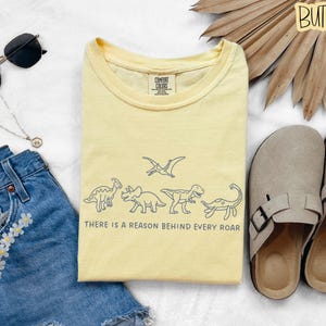 May include: A pale yellow t-shirt featuring a dark blue line art graphic of five dinosaurs and a pterodactyl, with the text "THERE IS A REASON BEHIND EVERY ROAR" printed below the illustrations. The shirt has a crew neckline and a Comfort Colors label.
