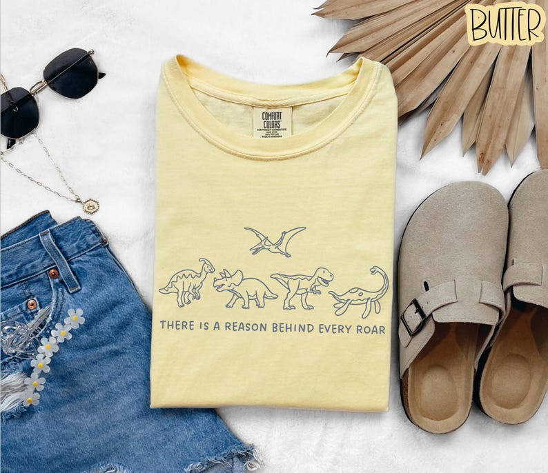 May include: A pale yellow t-shirt featuring a dark blue line art graphic of five dinosaurs and a pterodactyl, with the text "THERE IS A REASON BEHIND EVERY ROAR" printed below the illustrations. The shirt has a crew neckline and a Comfort Colors label.