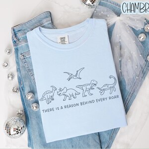 May include: A light blue t-shirt featuring a dark blue line art illustration of four dinosaurs and a pterosaur. Below the dinosaurs, the text reads, "THERE IS A REASON BEHIND EVERY ROAR." The shirt is layered over blue denim jeans and a white tulle bow.