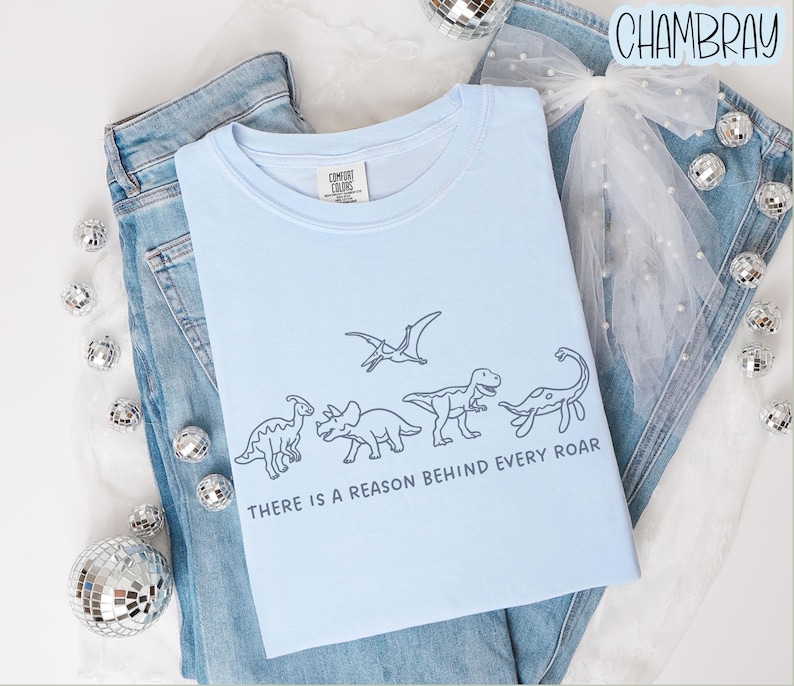 May include: A light blue t-shirt featuring a dark blue line art illustration of four dinosaurs and a pterosaur. Below the dinosaurs, the text reads, "THERE IS A REASON BEHIND EVERY ROAR." The shirt is layered over blue denim jeans and a white tulle bow.
