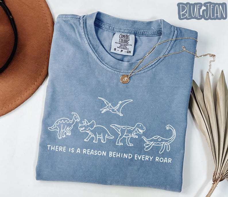 May include: A blue denim-colored t-shirt featuring a white line-art graphic of four dinosaurs and a pterodactyl. Below the dinosaurs, the text reads, "THERE IS A REASON BEHIND EVERY ROAR." The shirt has a Comfort Colors brand label at the neck.