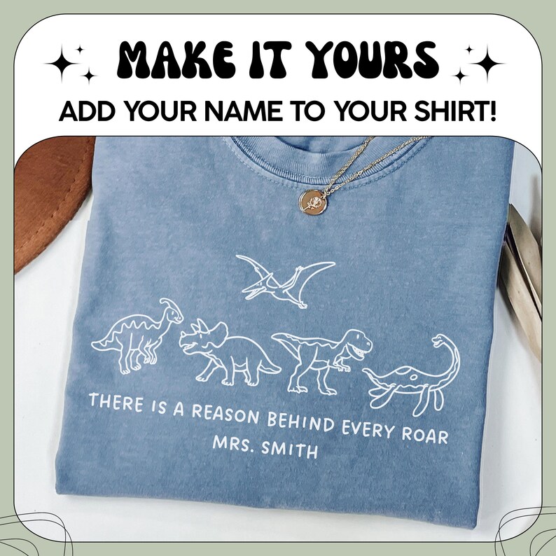 May include: A blue t-shirt featuring white line art of four dinosaurs and the text "THERE IS A REASON BEHIND EVERY ROAR MRS. SMITH". Above the shirt, the text reads "MAKE IT YOURS" and "ADD YOUR NAME TO YOUR SHIRT!" with star icons.