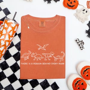May include: A rust-colored short-sleeve t-shirt featuring a white line-art graphic of four dinosaurs and a pterodactyl. Below the dinosaurs, the text reads, "THERE IS A REASON BEHIND EVERY ROAR." The shirt has a crew neckline and a size XL label.