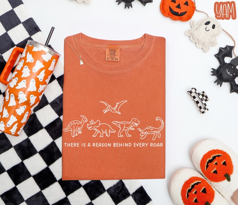 May include: A rust-colored short-sleeve t-shirt featuring a white line-art graphic of four dinosaurs and a pterodactyl. Below the dinosaurs, the text reads, "THERE IS A REASON BEHIND EVERY ROAR." The shirt has a crew neckline and a size XL label.