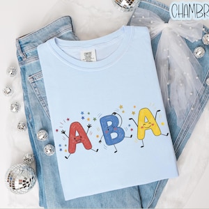 May include: A light blue t-shirt featuring a graphic of the letters A, B, and A as animated characters with arms and legs, surrounded by small colorful stars. The shirt is layered over blue denim jeans and a white sheer bow with pearl accents.