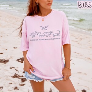 May include: A light pink short-sleeved t-shirt featuring a dark blue line art graphic of four dinosaurs and a pterosaur. Below the illustrations, the text reads, "THERE IS A REASON BEHIND EVERY ROAR." The shirt has a crew neckline and a loose fit.