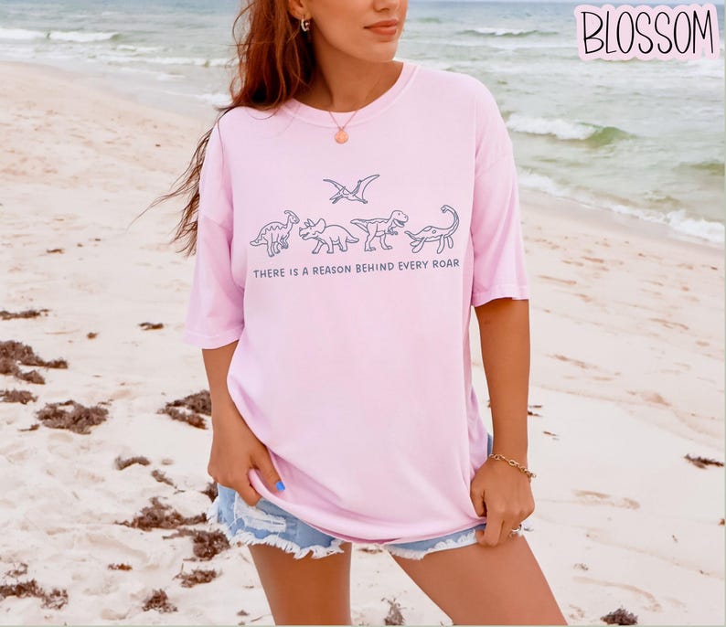 May include: A light pink short-sleeved t-shirt featuring a dark blue line art graphic of four dinosaurs and a pterosaur. Below the illustrations, the text reads, "THERE IS A REASON BEHIND EVERY ROAR." The shirt has a crew neckline and a loose fit.