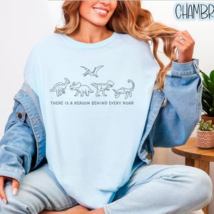 May include: A light blue crewneck t-shirt featuring a black line art graphic of four dinosaurs and a pterosaur, with the text "THERE IS A REASON BEHIND EVERY ROAR" printed in a simple sans-serif font centered below the dinosaur illustrations.