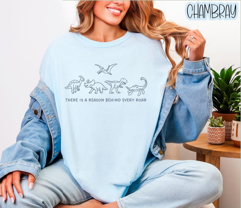 May include: A light blue crewneck t-shirt featuring a black line art graphic of four dinosaurs and a pterosaur, with the text "THERE IS A REASON BEHIND EVERY ROAR" printed in a simple sans-serif font centered below the dinosaur illustrations.