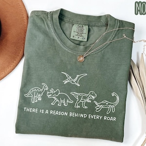May include: A moss green crew neck t-shirt featuring a white line art graphic of four dinosaurs and a pterodactyl. Below the dinosaurs, the text reads, "THERE IS A REASON BEHIND EVERY ROAR." The shirt has a Comfort Colors brand label at the neck.