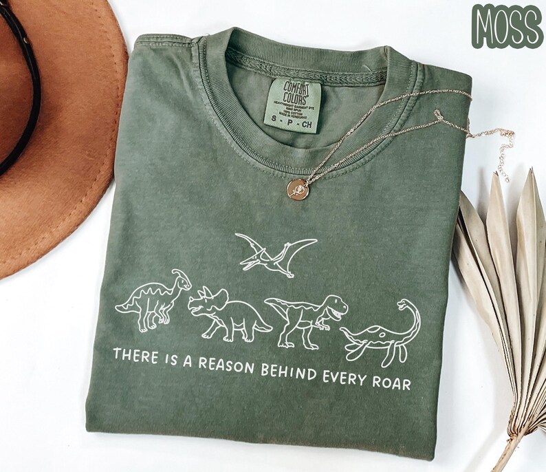 May include: A moss green crew neck t-shirt featuring a white line art graphic of four dinosaurs and a pterodactyl. Below the dinosaurs, the text reads, "THERE IS A REASON BEHIND EVERY ROAR." The shirt has a Comfort Colors brand label at the neck.