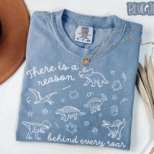 May include: A blue short-sleeved t-shirt featuring white line art of various dinosaurs, puzzle pieces, and stars. The text reads, "There is a reason behind every roar." The shirt has a Comfort Colors brand label at the neck and a size S-P-CH tag.