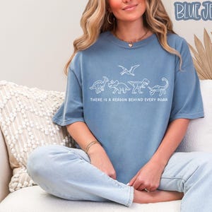May include: A blue denim-colored t-shirt featuring a white line-art graphic of four dinosaurs and a pterosaur. Below the illustration, the text reads, "THERE IS A REASON BEHIND EVERY ROAR." The shirt has a crew neckline and short sleeves.