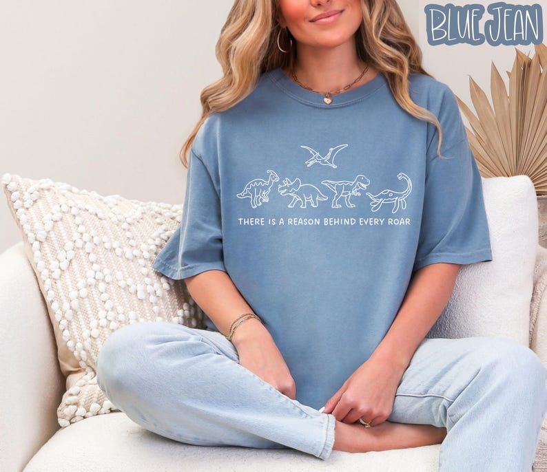 May include: A blue denim-colored t-shirt featuring a white line-art graphic of four dinosaurs and a pterosaur. Below the illustration, the text reads, "THERE IS A REASON BEHIND EVERY ROAR." The shirt has a crew neckline and short sleeves.